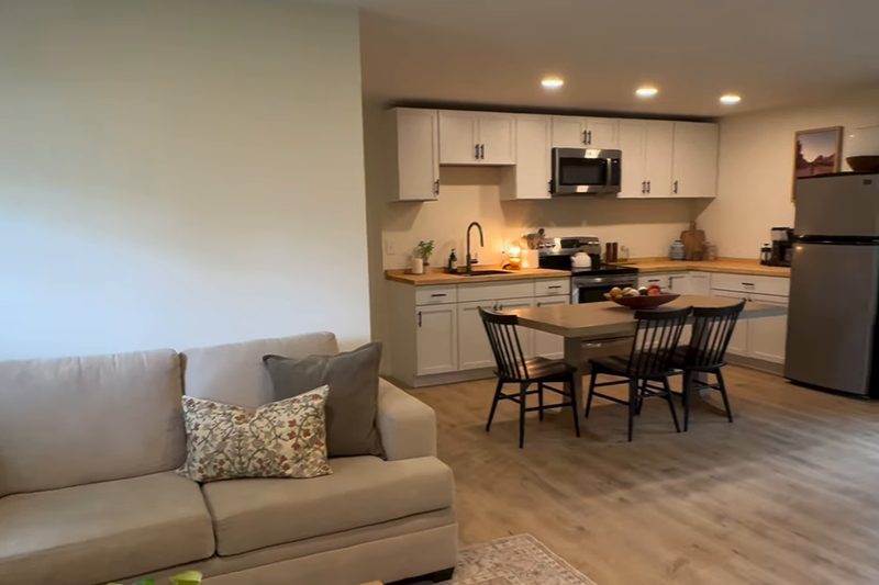 Finished home addition with an open living area, sofa, rug and coffee table, and a kitchen with white cabinets and a dining table beyond